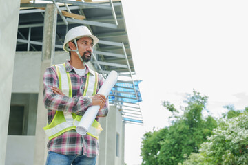 Asian male architect standing outdoors at a construction site, holding rolled-up blueprints....