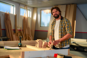 Portrait of young bearded hipster woodworker who sanding a piece of wood in workshop.