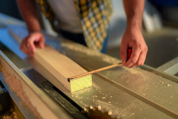 Close up hands of woodcrafter measuring a wooden plank with a tape measure on big table saw in a workshop.