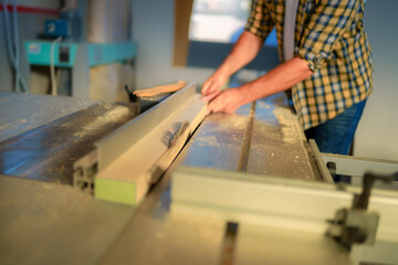 Close up on hands of woodworker in a workshop, using a table saw to cut board. He is wearing safety glasses and a checkered shirt.