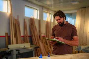 Bearded carpenter dressed in work clothes checking notes and making sketches on a clipboard.