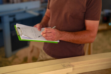 Craftsmen is working in a woodworking shop. He is focused on examine and take notes on paper plans for crafting, surrounded by wooden materials and tools.