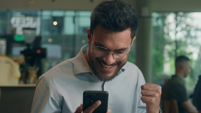 Male customer visitor tourist in cafe restaurant happy cheerful amazing Indian Arabian businessman ethnic business man guy worker read mobile phone message rejoice celebrate win online bet gambling
