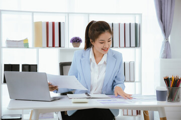 Confident Asian businesswoman in modern office smiling while working on laptop, analyzing reports, planning marketing strategies and leading with energy and positivity.