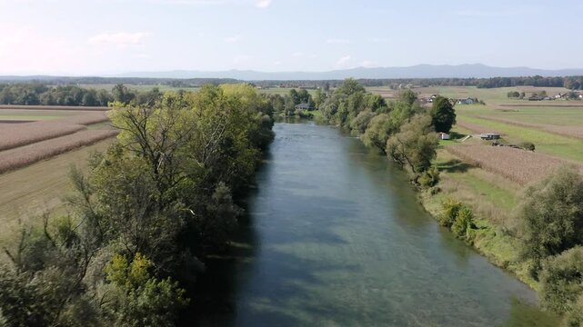 Flying above calm river Kolpa flowing gently between thick clusters of trees. A small rural house sits along the riverbank, surrounded by greenery and peaceful countryside.