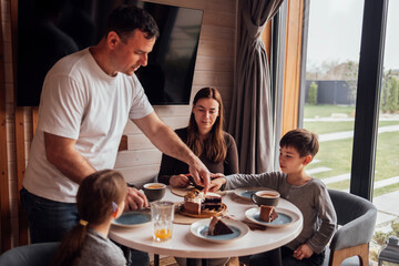 Happy family is having lunch in dining room at home. Man laughs and cuts pie. Dad, mom and their cute children are having breakfast together.