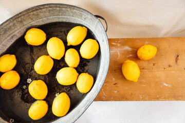 Juicy fresh lemons and daisies in water in a vintage metal basin. Top view. Wooden retro bench with yellow citrus fruits. White fabric in the background.