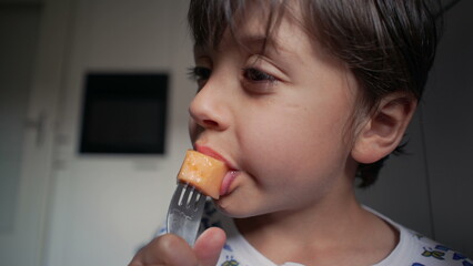 Close-up of young boy eating a piece of cantaloupe with a fork, highlighting the enjoyment of a...