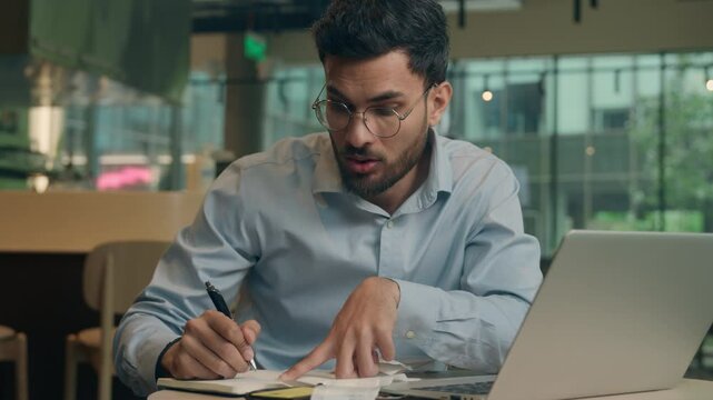 Indian man male Arabian guy businessman freelancer manager business entrepreneur work online in cafe restaurant write paperwork notes count bills writing notebook confused shock debt credit payment