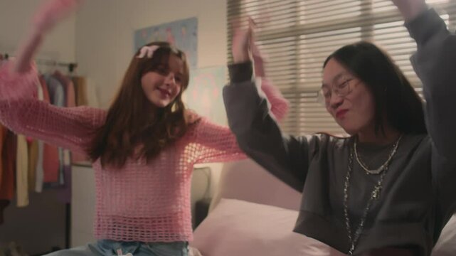 Two multi-ethnic teenage best friends smiling and rehearsing k-pop dance while sitting on bed
