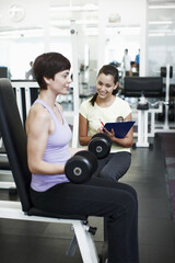 Fototapeta premium Personal trainer, woman and dumbbell at gym with clipboard, notes and assessment for progress. People, man and coach with checklist for stats, fitness and exercise with target at wellness club