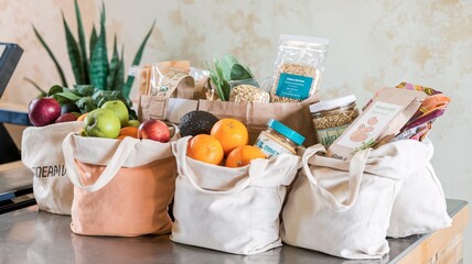 A close-up of reusable cloth bags filled with fresh produce, grains, and eco-friendly packaged goods at the checkout counter. The texture of the bags and fresh ingredients is highlighted.