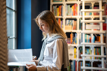 Immersed in studies middle aged student woman reading textbook standing near window in university library. Interested focused female shopping in bookstore choosing book. Literature lover in bookshop.