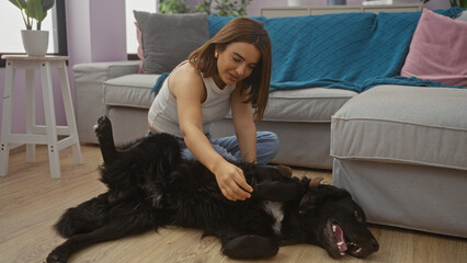 A young hispanic woman smiling and playing with her black labrador dog indoors in her living room, featuring cozy furniture and bright decor.