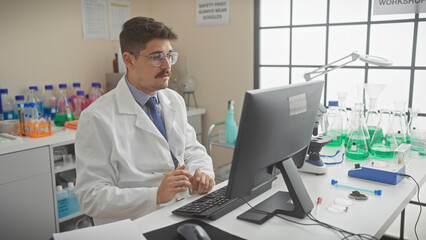 Young hispanic scientist in white lab coat working on computer in modern laboratory setting