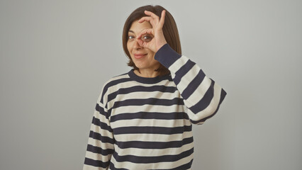 Hispanic middle-aged woman making an okay gesture near her eye against a white background, wearing a striped shirt.