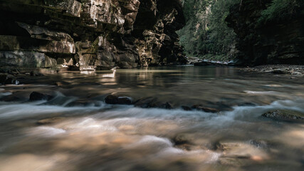 A vibrant stream rushes through a forested gorge, bouncing over rocks and creating a dynamic, lively scene. The surrounding trees and rocky terrain frame the water perfectly.