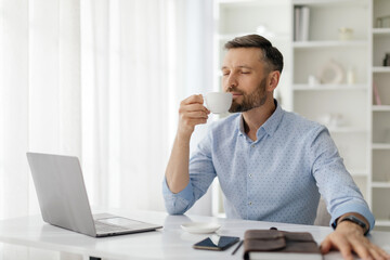 A man with a beard sits at a desk, savoring a cup of coffee. He is focused on his laptop, immersed...