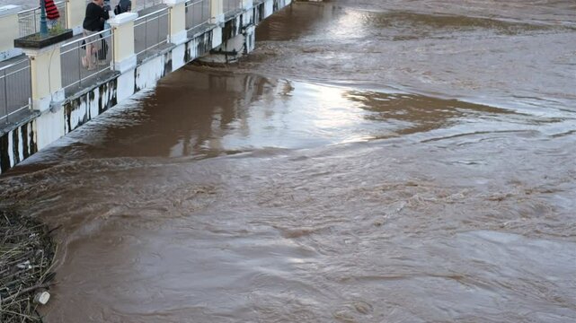 A river is flooded with water and people are standing on a bridge. Scene is one of concern and worry, as the people are likely trying to avoid the floodwaters and stay safe