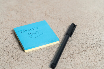 Small blue notepad on stone table with word thank you written by hand next to pen