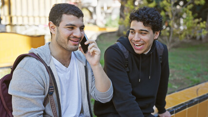 Two smiling hispanic men in casual attire enjoying a sunny day outdoors in the park, with one talking on a cellphone.