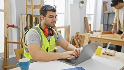 Two men work in a bright carpentry workshop, one using a laptop while the other measures wooden planks.