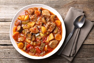 Delicious stew with vegetables in bowl and spoon on wooden table, top view