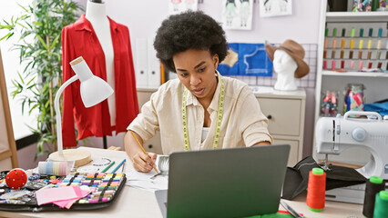 A focused black woman tailoring in a modern atelier, with sewing equipment and a laptop.