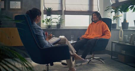 Obraz premium Back view of female therapist with notebook sitting in chair, providing therapy for teen boy. African American teenager shares feelings, discusses problems with psychologist during counseling session.
