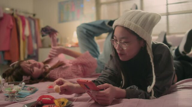 Korean teenager in white hat relaxing on bed holding smartphone, eating lychee and listening to k-pop music with her Caucasian girl friend