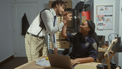 A smiling policewoman and her colleague celebrate with a high-five in their office, surrounded by...