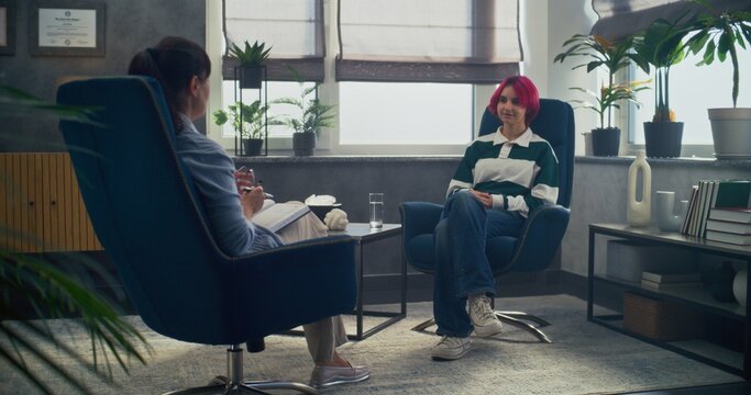 In psychotherapy room, teenage girl talks to her therapist, shares positive emotions during counseling session. Back view of female psychologist sitting in chair, providing therapy for to teenager. - Powered by Adobe