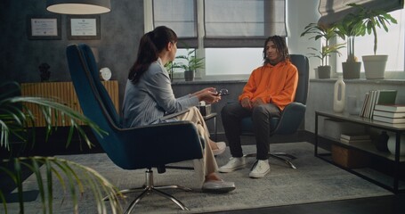 Female therapist sits in chair, takes notes in notebook. African American teen boy comes in psychological office on counseling session. Professional psychologist starts therapy, talks to teenager. © Framestock