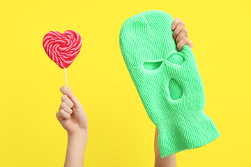 Female hands with balaclava and lollipop on yellow background, closeup