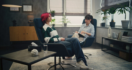 Girl and her psychologist in psychotherapist's office. Young patient sitting with her leg over knee, talking about experience and sharing positive emotions. Female therapist watches and listens.