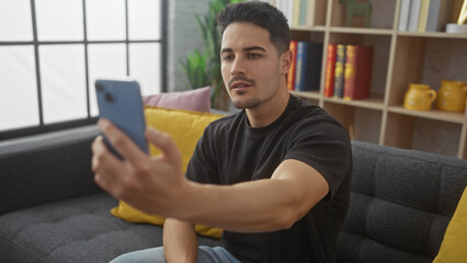 Handsome young hispanic man with a beard taking a selfie in his cozy living room.