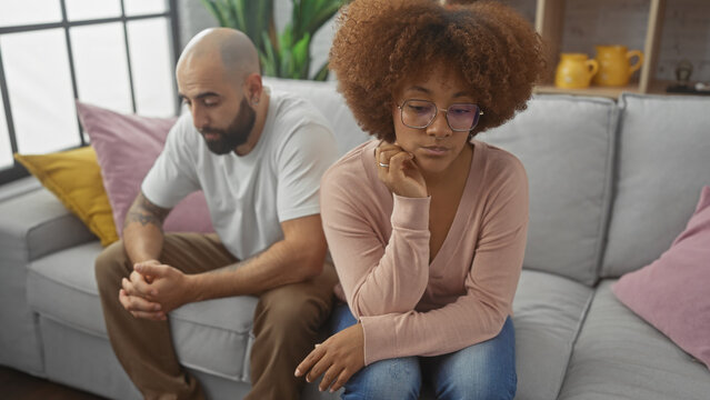 A thoughtful interracial couple sitting apart on a grey sofa, displaying tension and contemplation in a modern apartment.