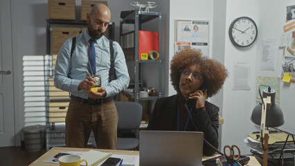 A woman talks on the phone while a man takes notes in a cluttered detective office space.