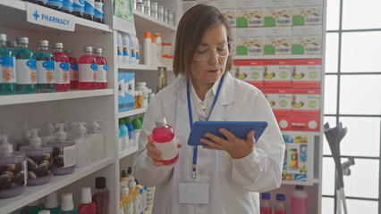 A middle-aged female pharmacist examines medication while using a tablet in a modern drugstore interior.