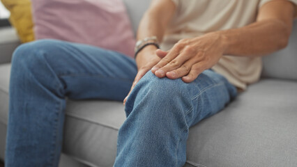 Hispanic man in pain holding his knee while sitting on a sofa indoors.