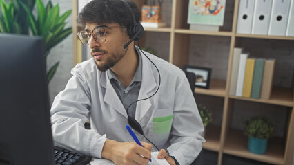 A young man with a beard in a clinic office wearing a headset and lab coat while working at a computer