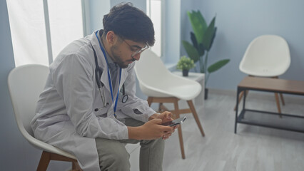 A young bearded man in medical attire using a smartphone in a modern clinic waiting room.