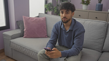 Fototapeta premium Handsome arab man with beard sitting in a modern apartment living room using smartphone