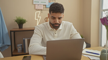 A focused hispanic man works on his laptop in a modern home office setting