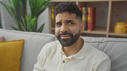 A young handsome hispanic man with a beard poses in a cozy home interior with a plant and bookshelf.