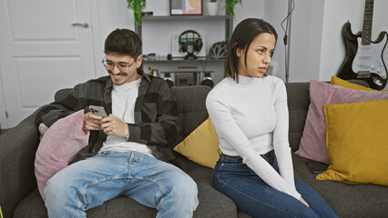 A woman looks concerned while a man smiles at his phone, sitting together on a couch in a cozy living room setting.