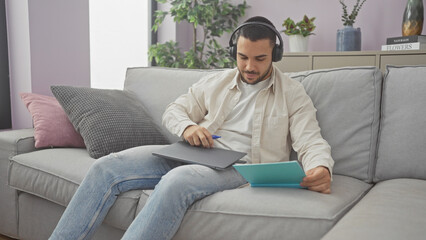 A young hispanic man dressed casually uses a graphic tablet while listening to music at home on a cozy sofa.