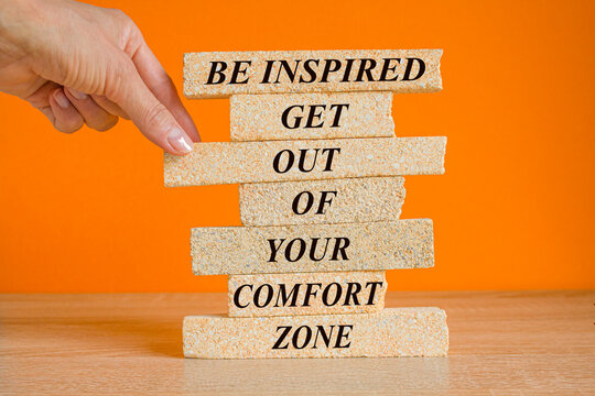 Motivational quote Be Inspired Get Out Of Your Comfort Zone written on brick blocks. Beautiful wooden table, orange background.