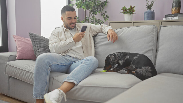A young hispanic man takes a photo of his teckel dog with his mobile phone while relaxing on a couch in a cozy home living room.