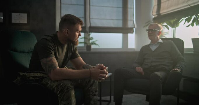Depressed military man with PTSD sits in chair, talks to specialist on counseling session. Senior psychotherapist listens and communicates with patient during therapy. Post traumatic stress disorder.